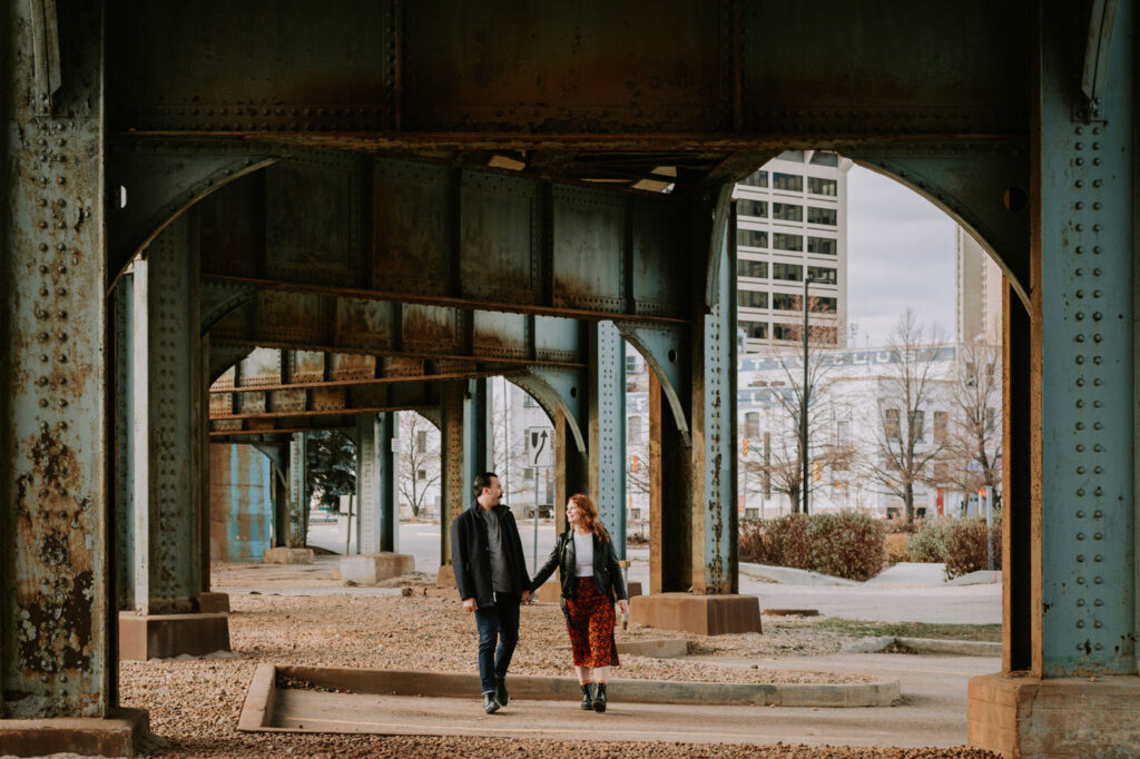 Exchange District engagement session in Winnipeg with couple holding hands below a train bridge